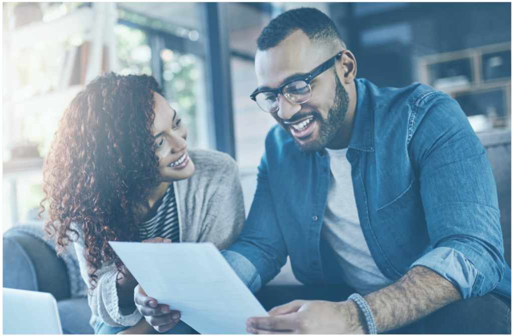 A man and woman sit on a couch looking at a piece of paper, smiling and engaged in conversation about their upcoming, low-cost colonoscopy.