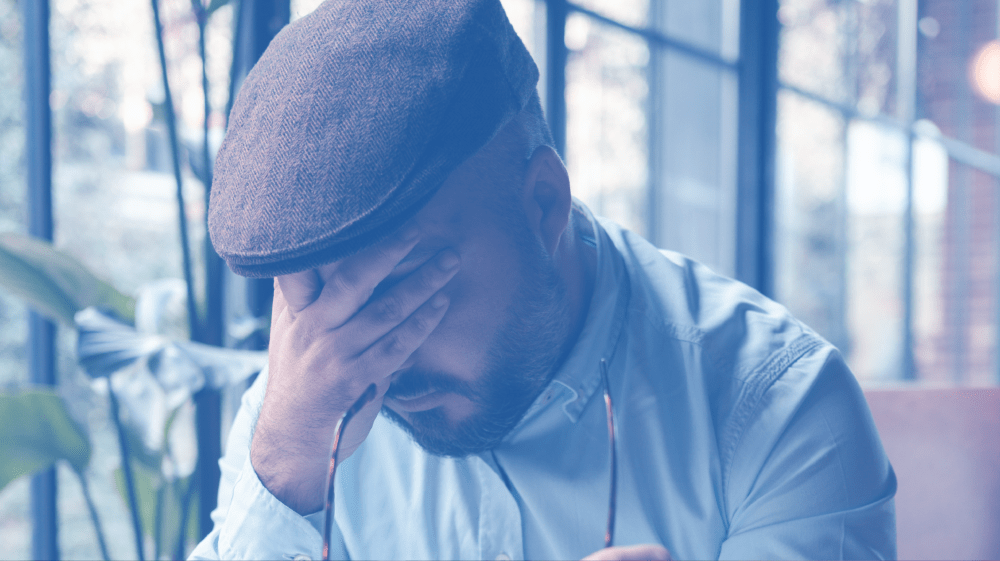 A man in a cap sits with hand covering his face inside a building with large windows, contemplating the journey ahead for his upcoming low-cost colonoscopy.