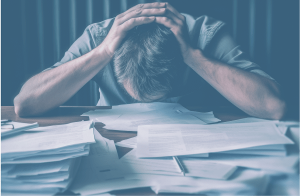 A person with head in hands sits at a desk piled with papers, appearing overwhelmed by the looming decision of scheduling a low-cost colonoscopy.