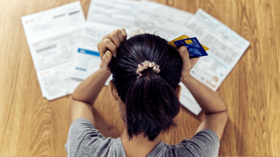 A person holding credit cards sits at a table covered with bills and documents, viewed from above.