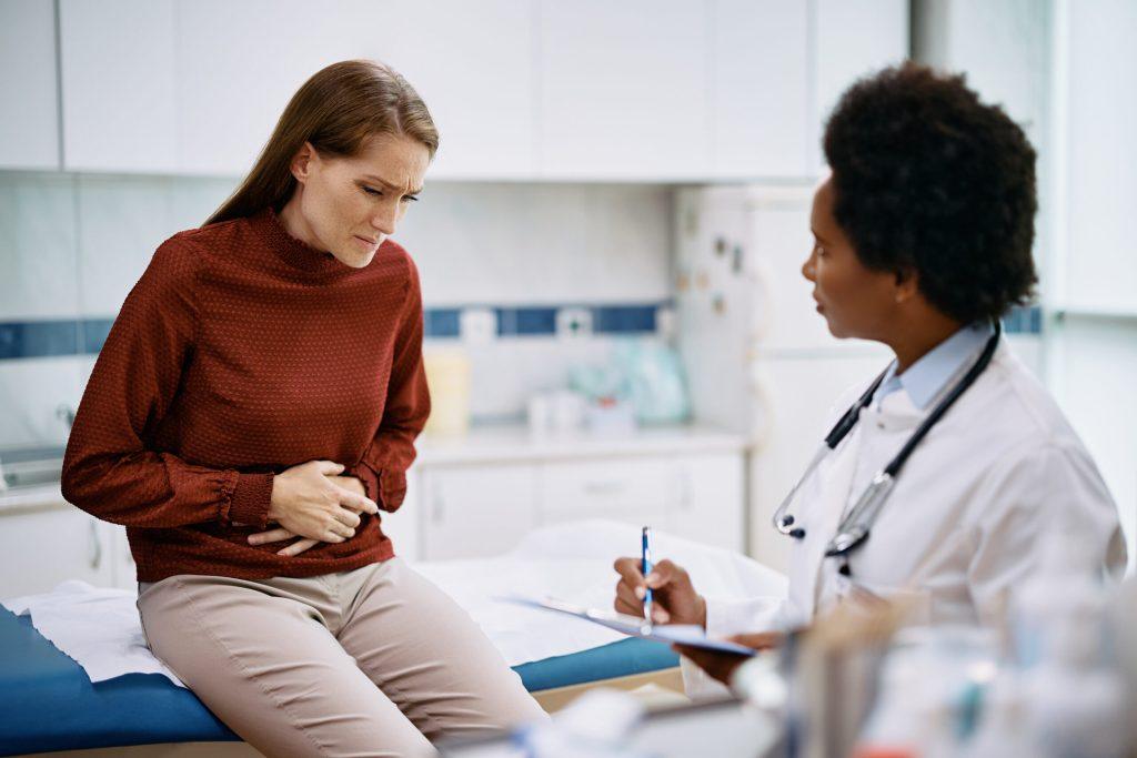 A woman holding her stomach in discomfort sits on an exam table while a doctor with a clipboard listens and takes notes, possibly discussing the need for a colonoscopy in the medical office.