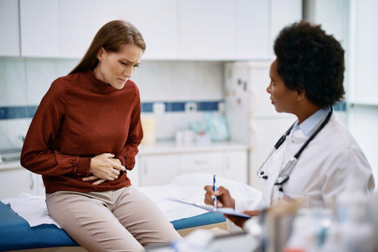 A woman holding her stomach in discomfort sits on an exam table while a doctor with a clipboard listens and takes notes, possibly discussing the need for a colonoscopy in the medical office.