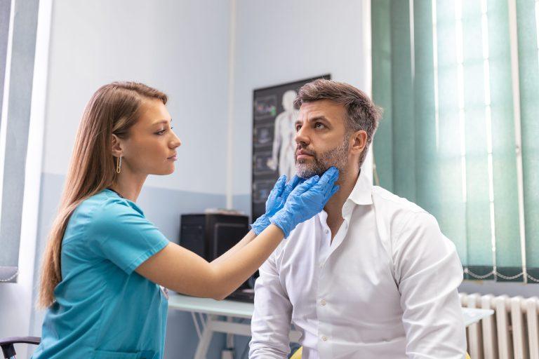 A healthcare professional wearing blue gloves examines a seated man’s neck and jaw area in a medical office, discussing his recent colonoscopy results.
