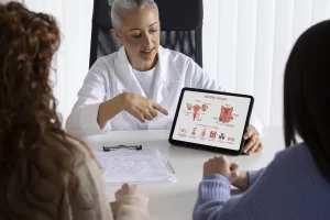 A doctor shows a tablet with female reproductive system diagrams, infertility causes, and discusses the possible role of colonoscopy during a consultation with two patients.