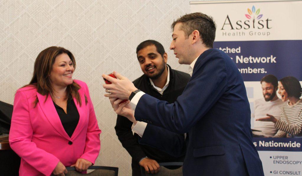 Three people converse near an Assist Health Group banner; one man in a blue suit shares colonoscopy information on his phone with the others, who are smiling.