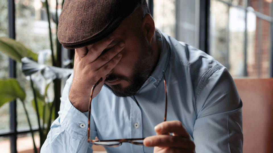 A person wearing a cap and light blue shirt sits indoors, holding glasses in one hand and covering their face with the other.