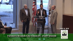 Three men stand and speak at a podium during a Farmers Branch City Council study session, discussing community health initiatives like colonoscopy awareness, with flags and a window in the background. The date shown is March 19, 2019.