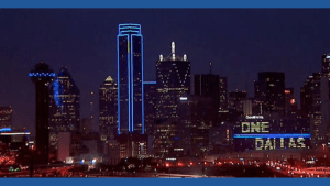 Dallas city skyline at night with illuminated buildings and a digital sign displaying “ONE DAY MAS, promoting awareness for colonoscopy screenings.