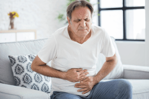 Older man sitting on a couch, holding his stomach with both hands and appearing to be in pain or discomfort, possibly awaiting a colonoscopy.