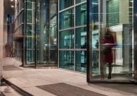 Glass-fronted office building entrance with revolving doors in use by blurred figures, suggesting movement and activity—much like the busy preparation ahead of a colonoscopy late at night.