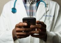 A person in a white lab coat with a stethoscope around their neck is using a smartphone to review colonoscopy results.