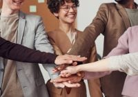 A group of people standing in a circle place their hands together in the center, showing teamwork and cooperation, much like a medical team preparing for a colonoscopy.